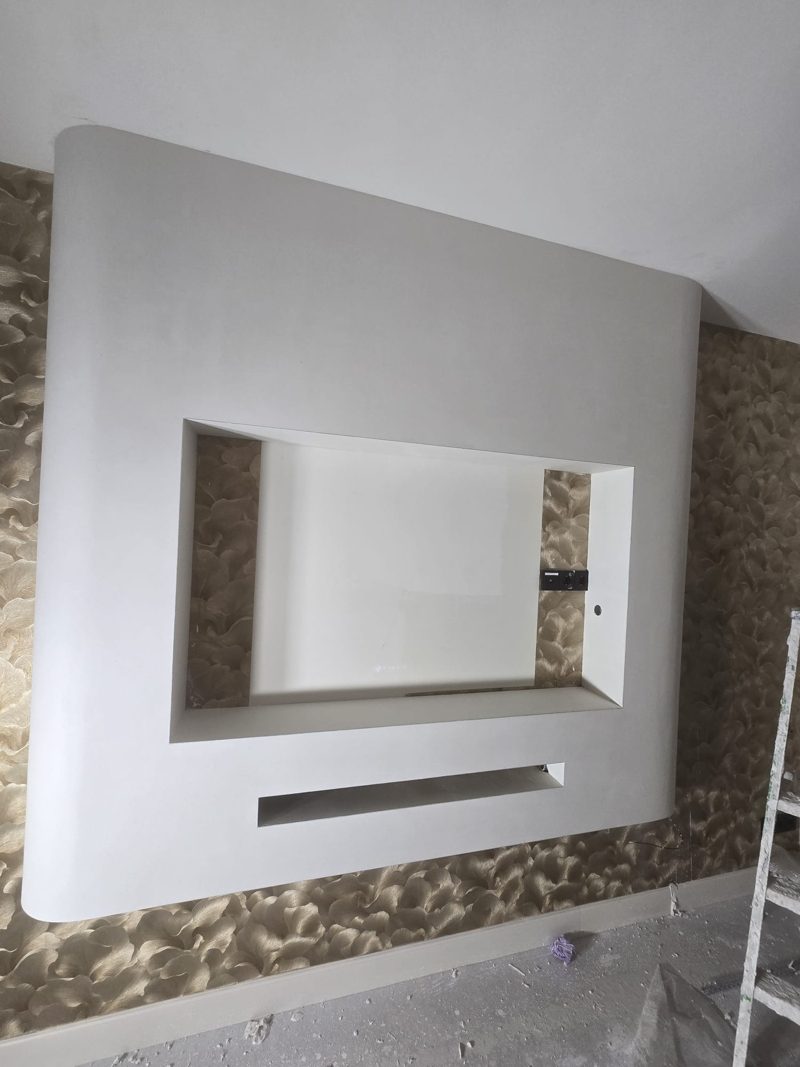 TV wall with rounded corners and a niche, plastered smooth and white, against a wall with gold-coloured wallpaper